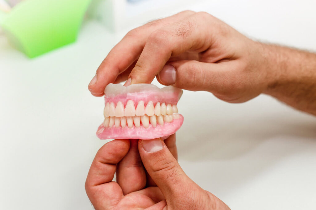 Mãos segurando modelo de prótese dentária. Solução para dentadura superior frouxa com prótese protocolo em Barreiras na Nakamura Odontologia. (143 caracteres)